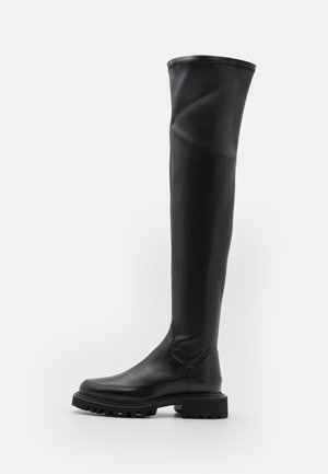 Botas negras por encima de la rodilla con material sintético suave, suela gruesa y puntera redondeada. Presentan un diseño sin costuras y un caña alta y flexible.