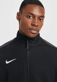 Zwarte zip-up jacket van soepel materiaal, met een hoge kraag en grijze accenten op de schouders. Logo zichtbaar op de linkerkant.