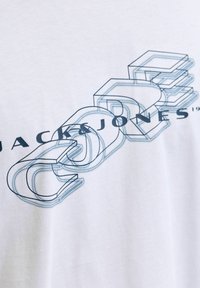 Bílý bavlněný tričko s modrou grafickou potiskem a textem "JACK & JONES" v stylizovaném, překrývajícím se písmu.