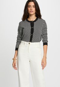 Cardigan à motifs noir et blanc avec un col rond, fermeture à boutons et poignets ajustés, porté avec un pantalon blanc taille haute.
