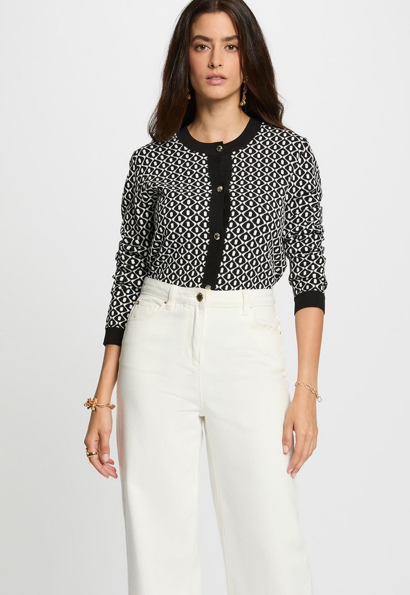 Cardigan à motifs noir et blanc avec un col rond, fermeture à boutons et poignets ajustés, porté avec un pantalon blanc taille haute.