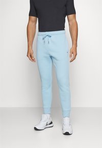 Calças de algodão azul claro com cintura elástica, cordão e punhos canelados. Combinadas com uma camisa preta de manga curta e sapatos brancos.