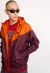 Cortavientos de Nike en naranja y burdeos, con capucha, puños elásticos y bolsillos con cremallera. Llevado sobre una camiseta gráfica roja.