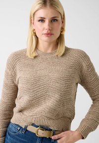 Pull beige en maille avec un motif chevron texturé, encolure ronde et ourlet côtelé. Associé à un jean bleu et une ceinture de couleur claire.