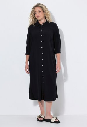 Blonde Frau mit lockigem Haar, die ein schwarzes midi-Kleid mit Knöpfen und dreiviertel-Ärmeln sowie schwarz-weiße flache Sandalen trägt.