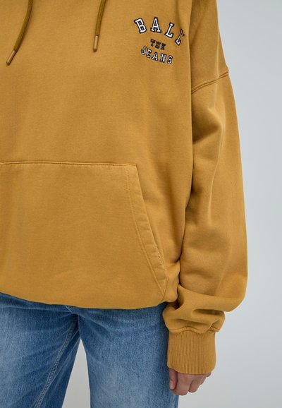 Dzeltena kapuce no kokvilnas, ar priekšējo kabatu un izšūtu logotipu. Piedurknes ir ribotas; valkāta ar gaiši ziliem džinsiem.