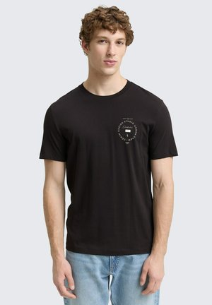 T-shirt noir à manches courtes en coton, avec un graphique circulaire blanc sur la zone gauche de la poitrine. Design ajusté, col rond.