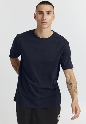 Tricou din bumbac bleumarin, cu guler rotund, mâneci scurte și textură netedă; purtat cu pantaloni închiși la culoare, având un detaliu cu logo.