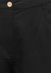 Primer plano de la cintura de unos pantalones negros con un botón beige y trabillas visibles, con detalles de costuras.