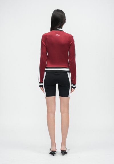 Červená sportovní bunda s černými a bílými pruhy, střih přiléhavý k tělu, doplněná černými cyklistickými kraťasy. Hladká textura, minimalistické logo na zadní straně.