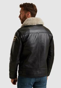 Črna usnjena jakna z kremasto ovčko, strukturna zasnova, hrbtne podrobnosti vključujejo strap hardware in vezenino.