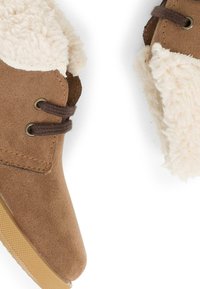 Botas de tobillo de ante marrón con un borde de borreguito crema y esponjoso, con cordones marrones y ojales en tono dorado, suela de goma texturizada.