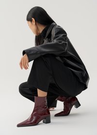 Bordeaux patent leren enkellaarsjes met een spitse neus, textuurafwerking en een chunky hak, gedragen met zwarte wijde broek en een leren jas.