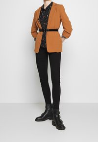 Orange blazer, sort blomster skjorte, sorte skinny bukser og sorte ankelstøvler med spænder. Glat stof, skræddersyet pasform, struktureret design.
