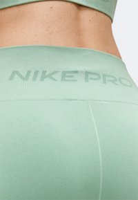 Κοντινό πλάνο στην κάτω πλάτη και τη μέση ενός ατόμου που φοράει ανοιχτοπράσινα κολάν Nike Pro με ραβδωτή μέση και ορατό λογότυπο.