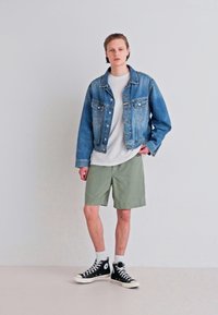Jachetă din denim cu detalii cu butoane, tricou alb, pantaloni scurți verde măsliniu, adidași negri cu glezna înaltă și șosete albe. Outfit simplu, casual.