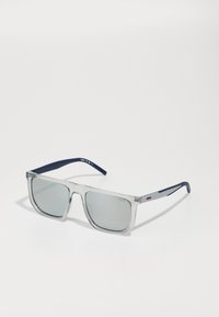 Ochelari de soare cu ramă din plastic transparent, lentile gri și brațe de culoare albastru marin, având un design minimalist și detalii subtile cu logo-ul mărcii.