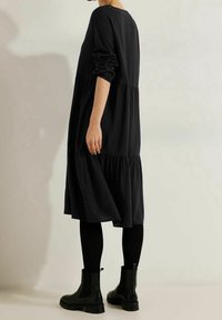 Schwarzes, schichtetes Kleid mit langen Ärmeln, aus weichem Stoff gefertigt. Hat eine lockere Passform und reicht bis unter das Knie. Kombiniert mit schwarzen Stiefeletten.