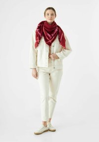 Femme se tenant debout portant une veste en denim écrue et un pantalon, des baskets blanches, et une grande écharpe rouge avec du texte enroulé autour du cou.