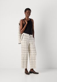 Donna con top senza maniche nero, pantaloni bianchi a scacchi, scarpe nere, che tiene una giacca abbinata sulla spalla contro uno sfondo semplice.