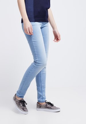 Jean skinny bleu clair en denim extensible associé à des baskets noires et blanches à imprimé serpent avec des lacets noirs. La jambe droite du mannequin est en avant.