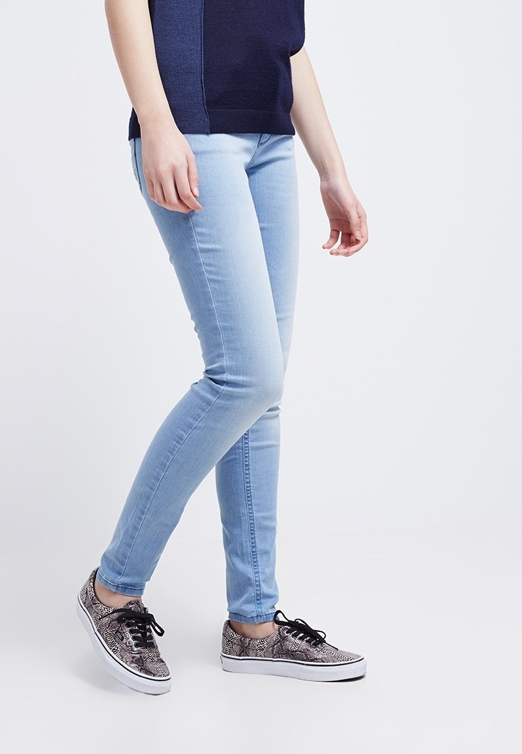 Jean skinny bleu clair en denim extensible associé à des baskets noires et blanches à imprimé serpent avec des lacets noirs. La jambe droite du mannequin est en avant.