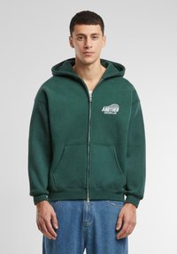 Zelena zip-up dukserica napravljena od pamuka, s kenguruzim džepom i otisnutim logotipom na prsima. Glatka tekstura i opušteni kroj.