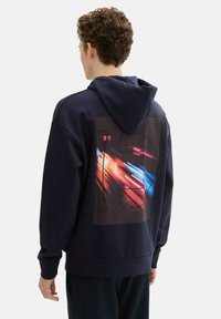 Granatowa bluza z kapturem z dużą, kolorową grafiką na plecach przedstawiającą abstrakcyjne smugi oraz numer "13". Miękki materiał, luźny krój.