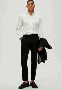 Camisa branca de botões com mangas longas, calças pretas slim fit e sapatos escuros; segurando um casaco preto numa mão. Design simples e moderno.