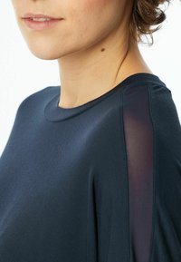 Navy blue top made from soft fabric, featuring a round neckline and sheer panels on the shoulders, shown from the side view.