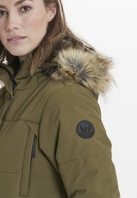 Olivgrön parkas med furtrim på huvan, fickor med dragkedja och svart logotypmärke på ärmen. Mjuk textur, skräddarsydd passform och hållbart tyg.