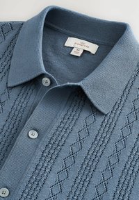 Jasnoniebieska dzianinowa koszulka polo z teksturowanym wzorem ściegu, zapięciem na guziki i krótkim kołnierzykiem. Posiada trzy szare guziki na listwie zapinającej.