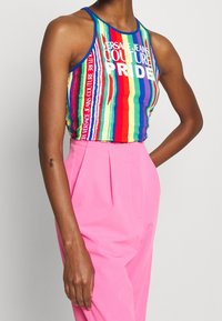 Personne portant un haut sans manches à rayures arc-en-ciel avec le texte "Versace Jeans Couture Pride" et un pantalon taille haute rose vif.