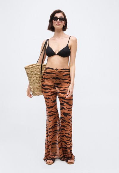 Γυναίκα με μαύρο τοπ μπικίνι, διάφανο παντελόνι φαρδύ με ρίγες τίγρης, κρατώντας μεγάλη υφαντή τσάντα και φοράει μαύρα σανδάλια και γυαλιά ηλίου.