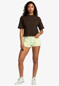 Ruskea ribattu crop-top, vihreät kukkakuosiset shortsit ja valkoiset korkeavartiset lenkkarit. Malli seisoo kädet taskuissa.