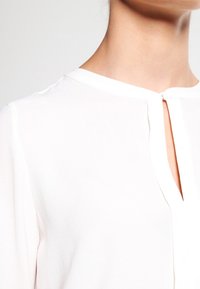 Blouse blanche en tissu lisse avec un col en V et un détail œillet à l'avant. Manches courtes et drapé subtil.