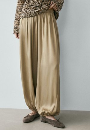 Pantaloni larghi color beige in raso con polsini elastici raccolti, indossati con ballerine marroni e un maglione lavorato a maglia grossa su una persona in piedi all'interno.