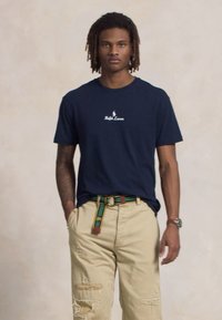 Jovem com dreadlocks, vestindo uma t-shirt azul marinho e umas calças bege desgastadas com um cinto riscado verde e amarelo, de pé com as mãos nos bolsos.