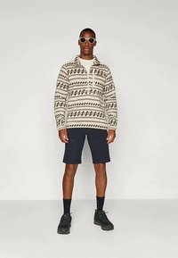 Πλεκτό πουλόβερ σε κρεμ απόχρωση με μαύρα γεωμετρικά σχέδια, με κολάρο που κλείνει με φερμουάρ, σε συνδυασμό με σκούρα σορτς και μαύρα αθλητικά παπούτσια. Το μοντέλο φοράει γυαλιά ηλίου.