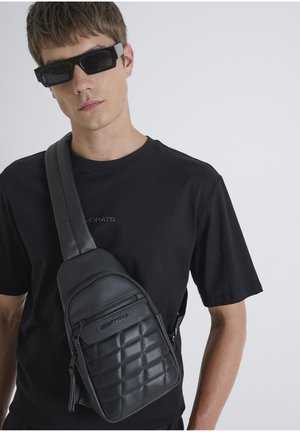 Bolso de hombro de cuero negro con un frente acolchado, doble cremallera y correa ajustable, llevado sobre una camiseta negra.