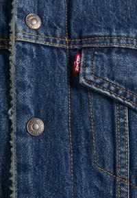 Veste en denim bleu avec surpiqûres orange, boutons métalliques et une petite étiquette rouge en tissu avec le logo Levi's sur la couture de la poche avant.
