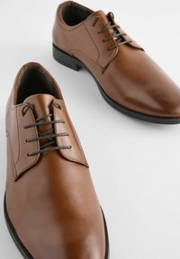 Chaussures en cuir marron avec une finition polie, bout rond et design à lacets avec cinq œillets. Présentent une semelle en caoutchouc noire et des détails de couture.