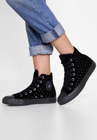 Svarta sammetshöga sneakers med nitar, rund tå och svarta gummisålar, tillsammans med uppvikta jeans.