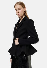 Czarny żakiet peplum o zarysowanej konstrukcji, z ostrymi klapami i akcentem w kolorze złota w postaci zapięcia. Teksturowany materiał dodaje głębi i elegancji.