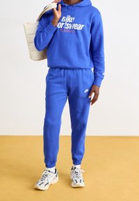 Blå hoodie med vit text "Nike Sportswear", matchande blå joggers och ljusa träningsskor med svarta accenter. Neutral bakgrund.