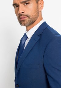 Abito blu navy con tessuto strutturato, rever a punta e una sola tasca sul petto. Abbinato a una camicia chiara e una cravatta blu con fantasia.