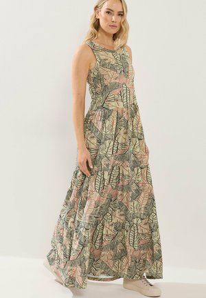 Vrouw in een mouwloze, tot op de vloer reikende jurk met een groene en roze tropische bladprint, staand tegen een effen witte achtergrond.