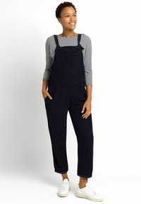 Marineblauwe corduroy salopette met een voorke pocket, gedragen over een gestreept lange mouwen shirt, met opgerolde mouwen en witte sneakers.