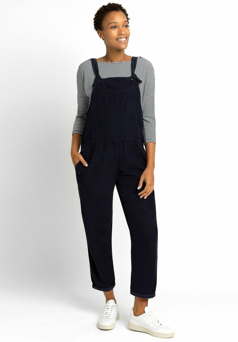 Marineblauwe corduroy salopette met een voorke pocket, gedragen over een gestreept lange mouwen shirt, met opgerolde mouwen en witte sneakers.
