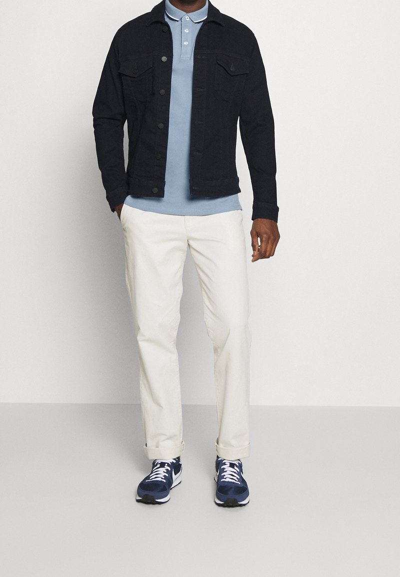 Homme portant une veste en jean noire, un polo bleu clair, un pantalon blanc et des baskets bleu marine, debout devant un fond uni.